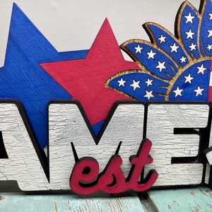 May include: Wooden sign with the word "Amer" in white letters with a distressed finish, "est" in red letters, and "1776" in white letters. The sign is decorated with a blue star, a red star, and a blue and white starburst.