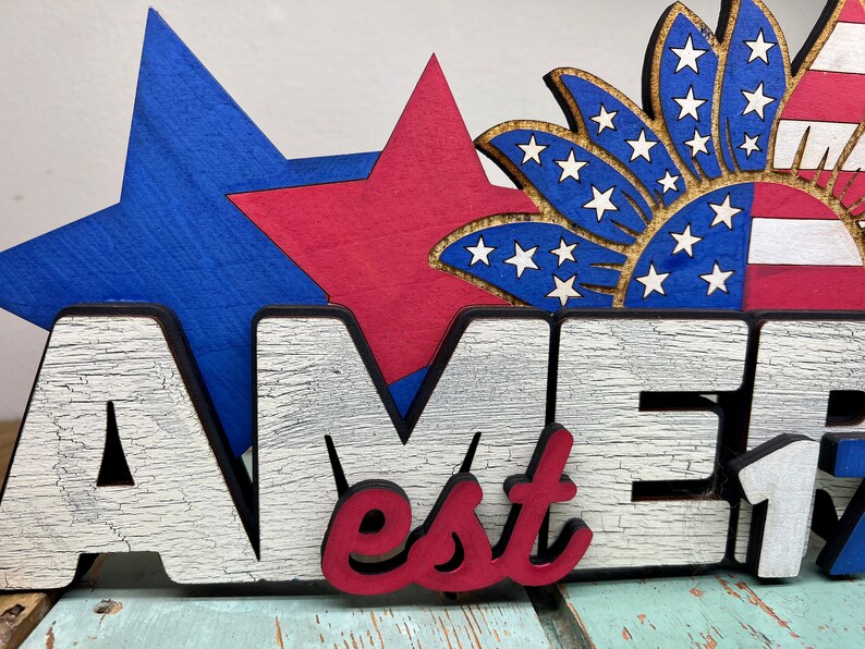 May include: Wooden sign with the word "Amer" in white letters with a distressed finish, "est" in red letters, and "1776" in white letters. The sign is decorated with a blue star, a red star, and a blue and white starburst.