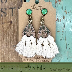 May include: A pair of wooden earrings with a green and gold leaf design and white tassel fringe. The earrings are decorated with green glitter and have a gold metal accent. The earrings are on a brown card with the word "Fall" written on it.