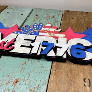 May include: Wooden sign with the words "America est 1776" in white and blue letters with a red, white, and blue sunflower design above the text.