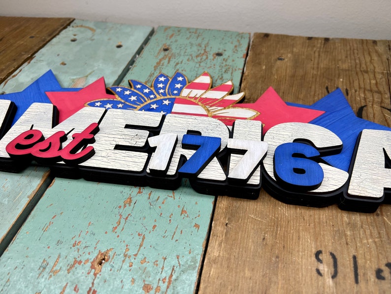 May include: Wooden sign with the words "America est 1776" in white and blue letters with a red, white, and blue sunflower design above the text.