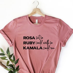 May include: A dusty rose t-shirt with black text that reads "ROSA sat so RUBY could walk so KAMALA could run".