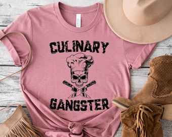 May include: A pink t-shirt with the text "CULINARY GANGSTER" in black. A skull wearing a chef's hat and crossed knives are below the text. The shirt is laid out on a wooden surface with a brown hat, a brown purse, and brown boots.