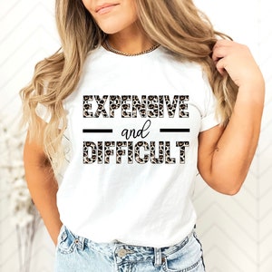 May include: White t-shirt with a leopard print graphic that says "EXPENSIVE and DIFFICULT" in black text.