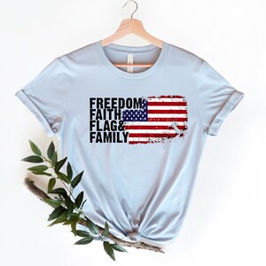 May include: Light blue t-shirt featuring a distressed American flag graphic and the words "Freedom, Faith, Flag & Family" printed in black.