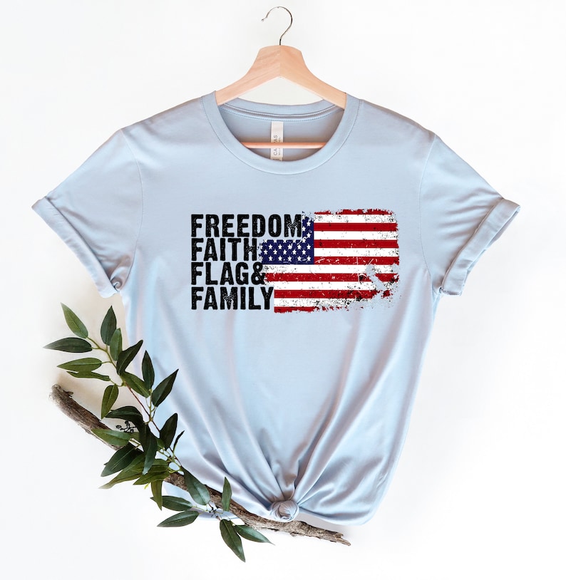 May include: Light blue t-shirt featuring a distressed American flag graphic and the words "Freedom, Faith, Flag & Family" printed in black.
