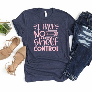 May include: A navy blue t-shirt with the text "I have no shelf control" in pink lettering. The text is accompanied by a stack of books. The shirt is displayed on a white background with a pair of brown sandals and a pair of blue jeans.