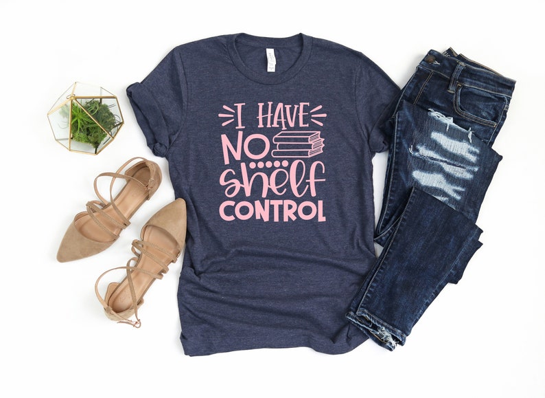 May include: A navy blue t-shirt with the text "I have no shelf control" in pink lettering. The text is accompanied by a stack of books. The shirt is displayed on a white background with a pair of brown sandals and a pair of blue jeans.