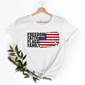May include: White t-shirt featuring a distressed American flag graphic and the words "Freedom, Faith, Flag & Family" printed in black.