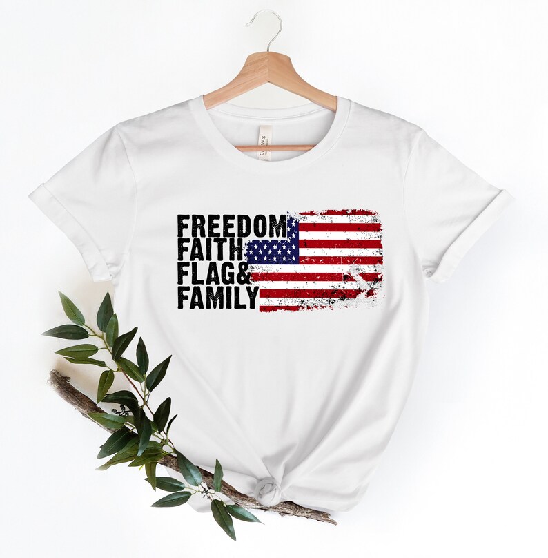 May include: White t-shirt featuring a distressed American flag graphic and the words "Freedom, Faith, Flag & Family" printed in black.