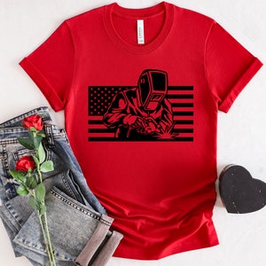 May include: Red t-shirt with a black silhouette of a welder in front of an American flag.