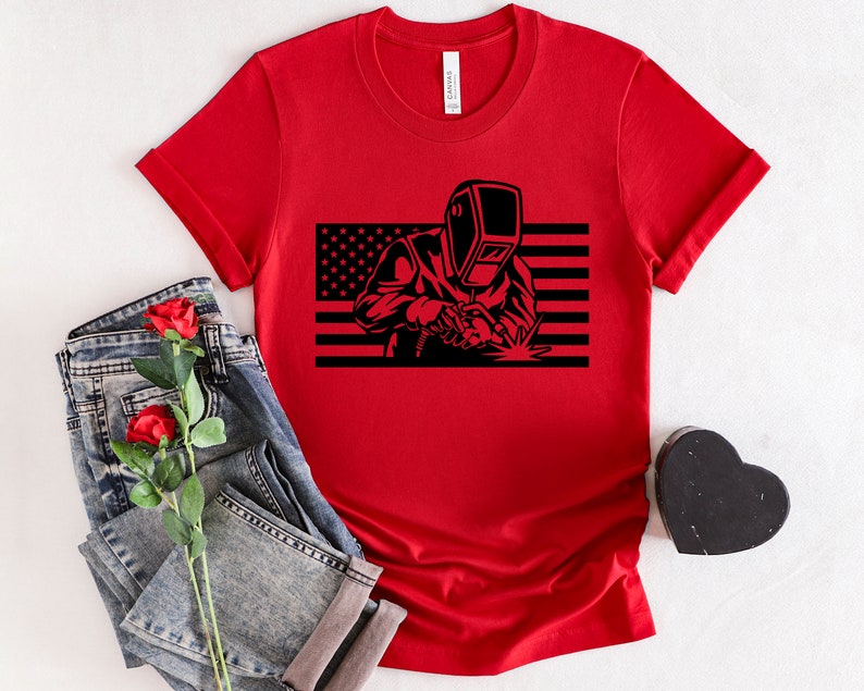 May include: Red t-shirt with a black silhouette of a welder in front of an American flag.