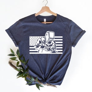 May include: A navy blue t-shirt with a white graphic of a welder wearing a welding mask in front of an American flag.