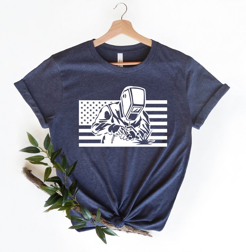 May include: A navy blue t-shirt with a white graphic of a welder wearing a welding mask in front of an American flag.