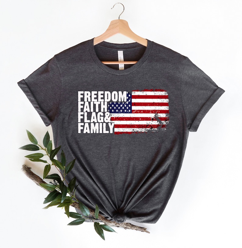 May include: A dark grey t-shirt featuring a distressed American flag design and the words "Freedom, Faith, Flag & Family" printed in white.