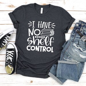 May include: A dark gray t-shirt with the text "I have no shelf control" in white. The text is accompanied by a stack of books. The shirt is laid out on a white wooden surface with a pair of black sneakers and a pair of blue jeans.