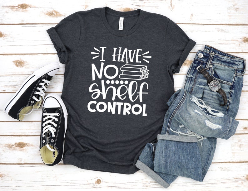 May include: A dark gray t-shirt with the text "I have no shelf control" in white. The text is accompanied by a stack of books. The shirt is laid out on a white wooden surface with a pair of black sneakers and a pair of blue jeans.