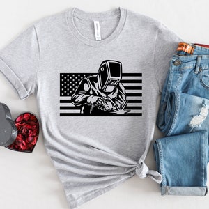 May include: A gray t-shirt with a black and white graphic of a welder wearing a welding mask in front of an American flag.