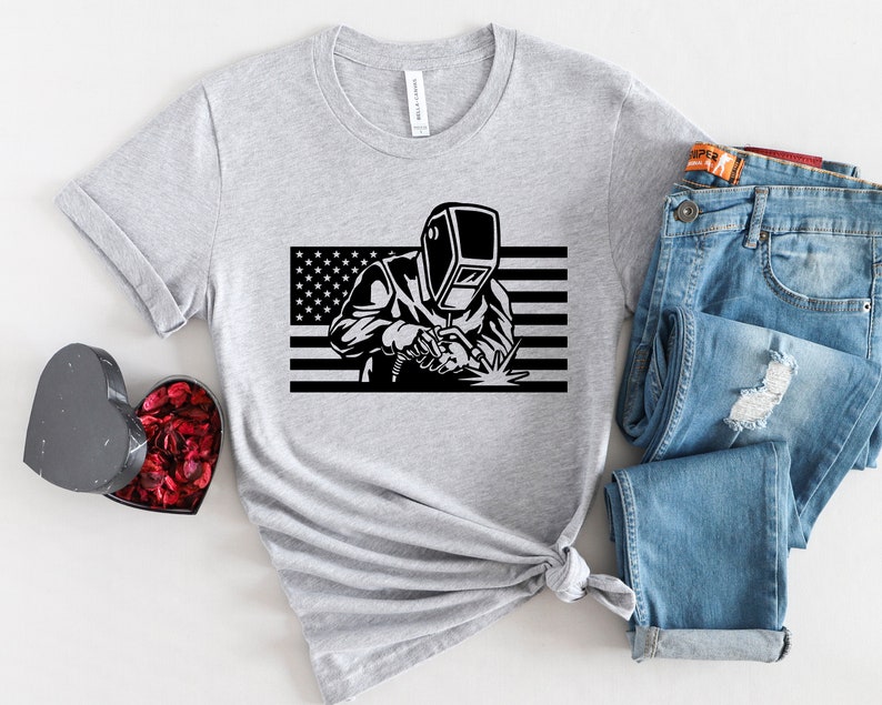 May include: A gray t-shirt with a black and white graphic of a welder wearing a welding mask in front of an American flag.