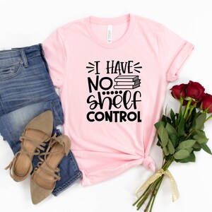 May include: A pink t-shirt with the text "I have no shelf control" and a stack of books. The shirt is laid out on a white background with a pair of brown sandals and blue jeans.