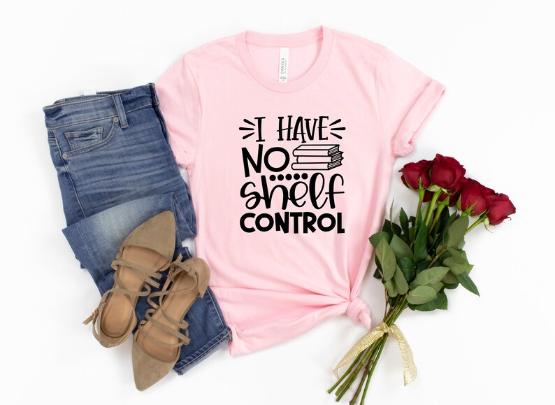 May include: A pink t-shirt with the text "I have no shelf control" and a stack of books. The shirt is laid out on a white background with a pair of brown sandals and blue jeans.