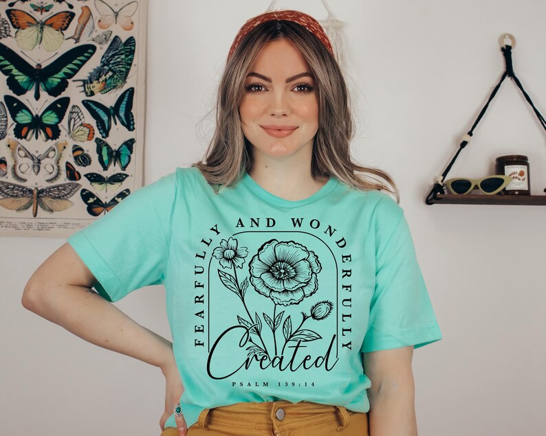 May include: A turquoise t-shirt with a black floral design and the text "Fearfully and Wonderfully Created" with the scripture reference "Psalm 139:14".