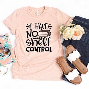 May include: A peach-colored t-shirt with black text that reads "I have no shelf control" with a stack of books above the word "no". The shirt is laid flat on a white background with a pair of white sandals, a pair of blue jeans, a floral arrangement, and a clutch purse.