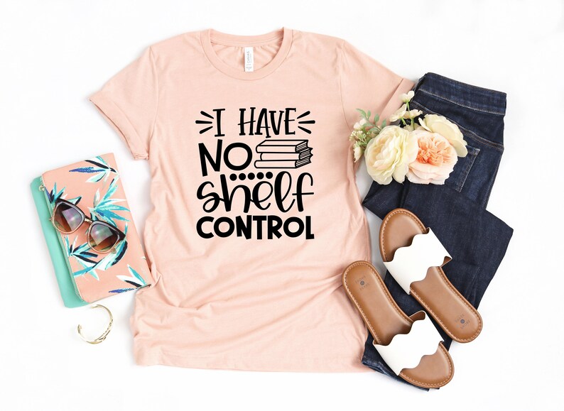 May include: A peach-colored t-shirt with black text that reads "I have no shelf control" with a stack of books above the word "no". The shirt is laid flat on a white background with a pair of white sandals, a pair of blue jeans, a floral arrangement, and a clutch purse.