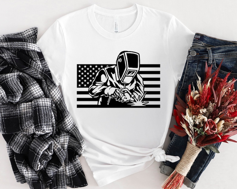 May include: White t-shirt with a black and white graphic of a welder wearing a welding mask in front of an American flag.