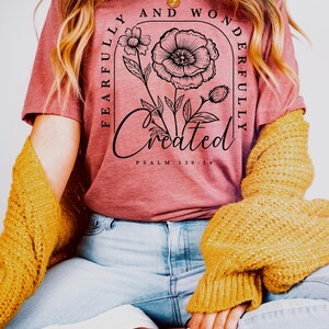 May include: A woman wearing a maroon t-shirt with a floral design and the text "Fearfully and Wonderfully Created Psalm 139:14".