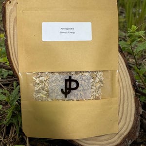 May include: A brown paper packet with a clear window, filled with dried herbs. The packet has a white label with the words "Ashwagandha" and "Stress & Energy". The packet is placed on a wooden slice.
