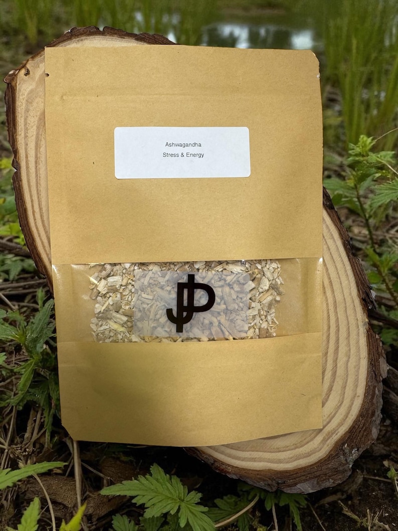 May include: A brown paper packet with a clear window, filled with dried herbs. The packet has a white label with the words "Ashwagandha" and "Stress & Energy". The packet is placed on a wooden slice.