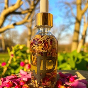 May include: A clear glass bottle with a gold cap and dropper, filled with dried flowers and petals. The bottle has a black logo. Pink flower petals and buds are scattered around the bottle, with a blurred background of trees and sky.