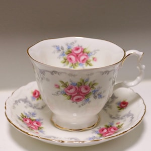 Puede incluir: Un juego de taza y platillo blancos con un diseño floral. La taza y el platillo están decorados con rosas rosas, detalles azules y ribetes dorados. La taza tiene un asa decorativa.
