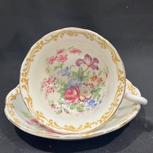 Może przedstawiać: Zestaw filiżanki i spodka do herbaty w stylu vintage. Biała porcelanowa filiżanka i spodek zdobione są kwiatowym wzorem z różowymi, fioletowymi, niebieskimi i żółtymi kwiatami. Złote zdobienia zdobią krawędzie obu elementów.
