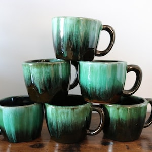 May include: Six ceramic mugs stacked in a pyramid formation. The mugs have a distinctive green and brown glaze, with dark brown handles. The mugs are a rounded shape with a glossy finish.