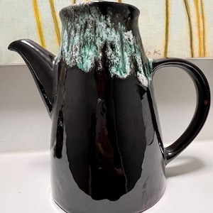 Op de afbeelding: Een zwarte keramische kan met een tuit en handvat. De bovenkant van de kan heeft een getextureerde, groen en witte glazuur. De kan wordt waarschijnlijk gebruikt voor het serveren van dranken.