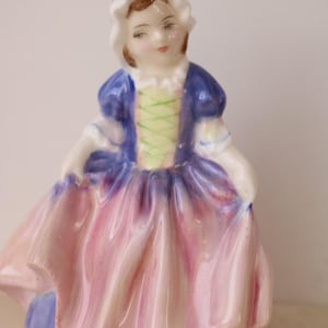 May include: A porcelain figurine of a woman in a historical dress. The figure wears a white bonnet, a blue bodice with a green laced front, and a pink and purple skirt. The figurine is a decorative item.