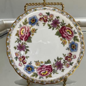May include: A small, round, white porcelain plate with a floral design. The plate features a scalloped edge with gold trim and a pattern of pink, blue, and purple flowers. The plate is displayed on a gold-colored metal stand.