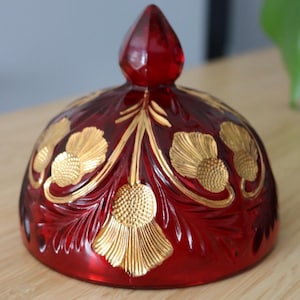 May include: A ruby red glass lid with a pointed finial. The lid is decorated with gold floral designs. The glass has a cut pattern, and the gold accents add a touch of elegance. This decorative piece is likely for a jar or container.