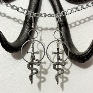 May include: Silver earrings with a circle design featuring a sword wrapped by a snake. The earrings are hanging from a silver chain.