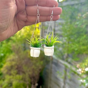 May include: A pair of silver dangle earrings featuring miniature white pots with green plants hanging from silver chains.