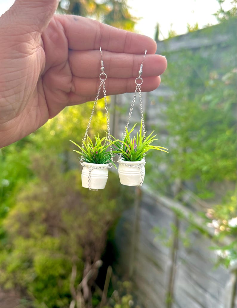 May include: A pair of silver dangle earrings featuring miniature white pots with green plants hanging from silver chains.