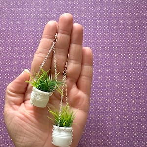 May include: A pair of dangle earrings featuring miniature white ceramic pots with green plants hanging from silver chains.