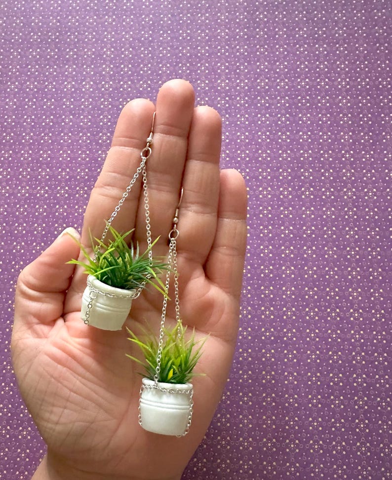 May include: A pair of dangle earrings featuring miniature white ceramic pots with green plants hanging from silver chains.