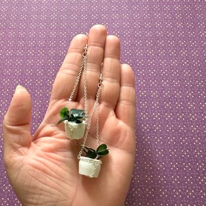 May include: A pair of silver dangle earrings featuring miniature white hanging planters with green plants.