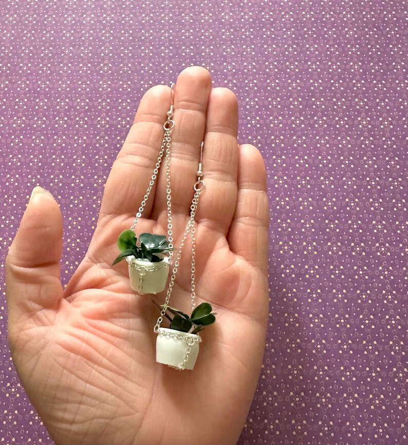 May include: A pair of silver dangle earrings featuring miniature white hanging planters with green plants.
