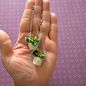 May include: A pair of silver dangle earrings featuring miniature hanging planters with green plants and yellow flowers.