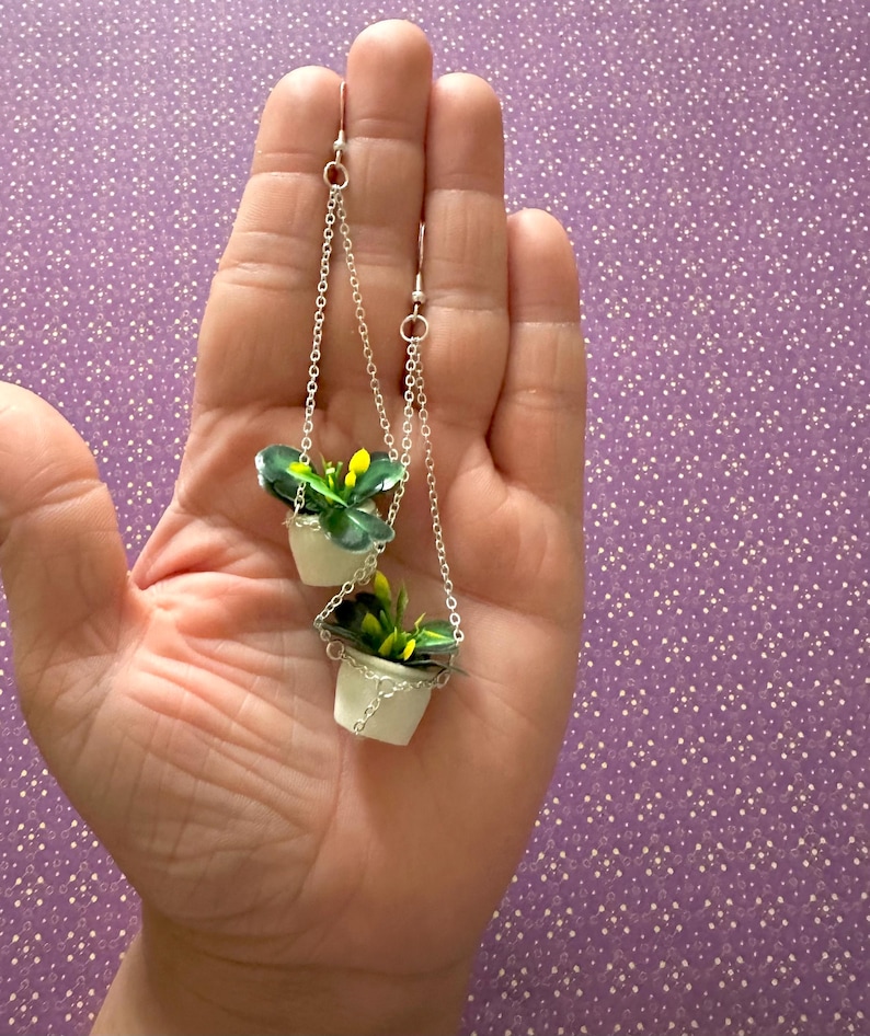 May include: A pair of silver dangle earrings featuring miniature hanging planters with green plants and yellow flowers.