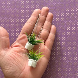 May include: A pair of silver dangle earrings featuring miniature white hanging planters with green plants.